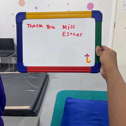Hand holding a small whiteboard with 'Thank you Miss Esther' written on it, against a colorful background.